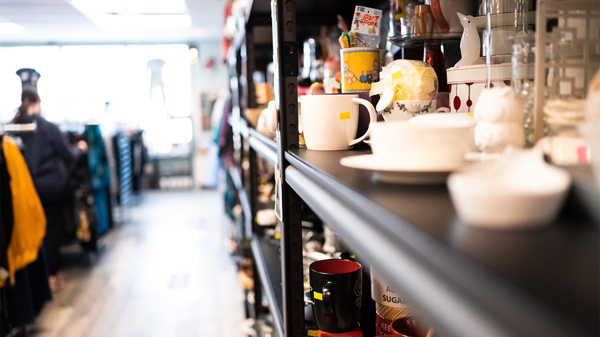 Teacups and glassware on black shelves in a thrift store, clothing racks in the background.