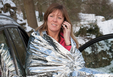 Women on phone leaning up against her car while wrapped in an emergency blanket to fend off the cold.